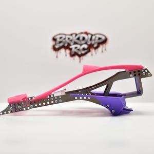 May include: A side profile of a radio-controlled car chassis featuring a dark metallic frame with numerous circular cutouts, a bright pink top rail, and a purple rear component. The logo BRKDUP RC is visible in the background.