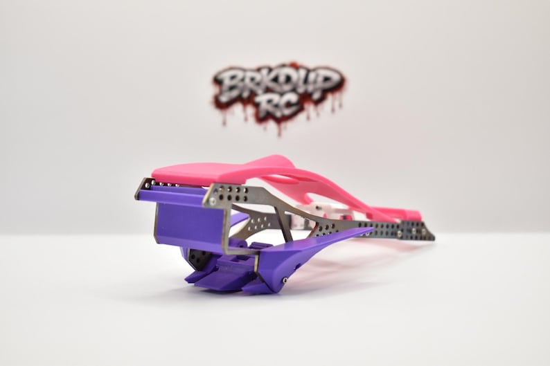 May include: A radio-controlled car chassis featuring a bright pink top section, a purple front bumper and lower components, and a dark grey metallic frame with perforated detailing. The logo BRKDP RC is visible in the background.