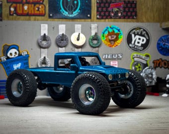 May include: A blue toy truck with large, black tyres. The truck has a flatbed and a small cab. A sticker on the back shows a skeleton in a shopping cart. The background includes stickers and metal objects.