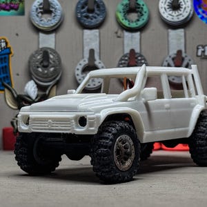 以下が含まれることがあります： 黒いオフロードタイヤを装着した白いおもちゃのSUV。車両にはロールケージとシュノーケルが付いています。背景には自動車部品と漫画のキャラクターが含まれています。