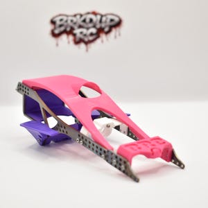 May include: A radio-controlled car chassis featuring a bright pink top panel, a purple rear section, and a white central component. The frame is constructed from dark grey perforated metal side rails. The logo BRKUP RC is visible in the background.