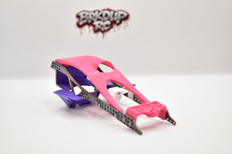 May include: A radio-controlled car chassis featuring a bright pink top panel, a purple rear section, and a white central component. The frame is constructed from dark grey perforated metal side rails. The logo BRKUP RC is visible in the background.