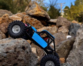 May include: A blue and black remote-controlled rock crawler vehicle with large, textured black tires and silver wheels. The vehicle features a skeletal frame design and is positioned at an incline on a large, porous brown rock surface.