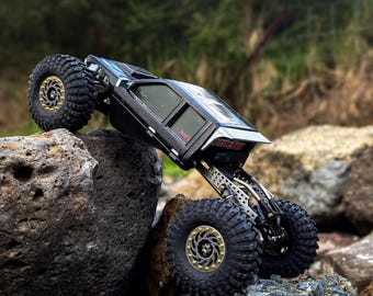 Peut inclure: Un véhicule télécommandé noir de type rock crawler avec de gros pneus à crampons dotés d'anneaux de blocage dorés. Le châssis est exposé avec des composants de suspension métalliques et la carrosserie affiche le texte Little Guy en rouge.