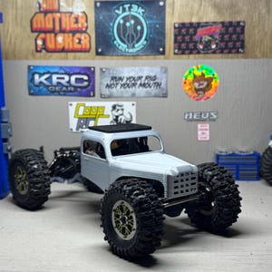 以下が含まれることがあります： 大きな黒いノブタイヤと詳細なシャーシを備えた、明るい灰色のRCトラック。トラックはクラシックなキャブデザインで、ガレージをテーマにした看板やステッカーを背景にしています。