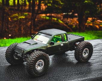 May include: A dark green toy truck with large, rugged tires. The truck has a black bed and a windshield with the text "#ROCKS". The background is blurred, showing green foliage and trees.