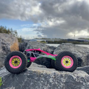 May include: A remote-controlled rock crawler vehicle featuring a bright pink and green chassis, black off-road tires, and neon pink wheel rims with yellow star-shaped centers, positioned on a large gray rock surface.
