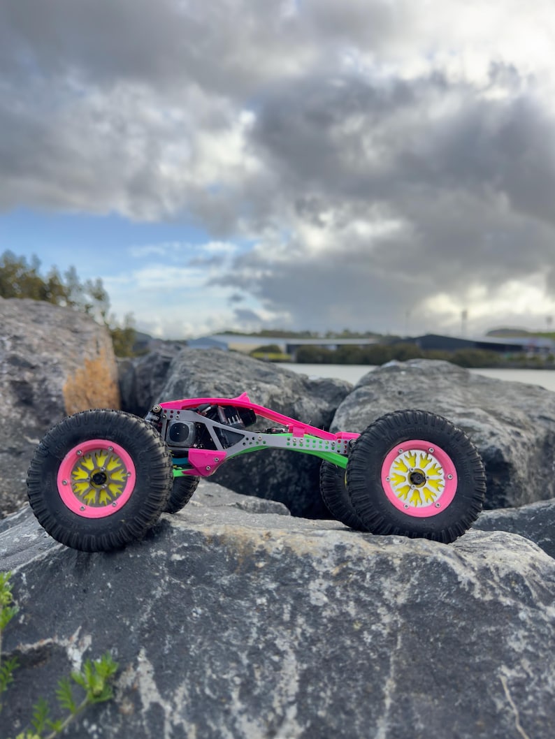 May include: A remote-controlled rock crawler vehicle featuring a bright pink and green chassis, black off-road tires, and neon pink wheel rims with yellow star-shaped centers, positioned on a large gray rock surface.