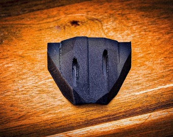 May include: A matte black, faceted, shield-shaped object with a textured surface. The item features a central vertical ridge and two elongated, recessed oval indentations positioned symmetrically on either side of the ridge, creating a geometric form.