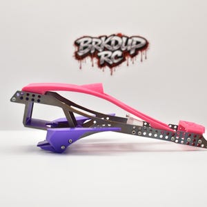 May include: A custom RC car chassis frame featuring a metallic grey perforated metal structure, a bright pink top section, and purple lower components. The logo BRKDUP RC is visible in the background in a stylized red and black font.