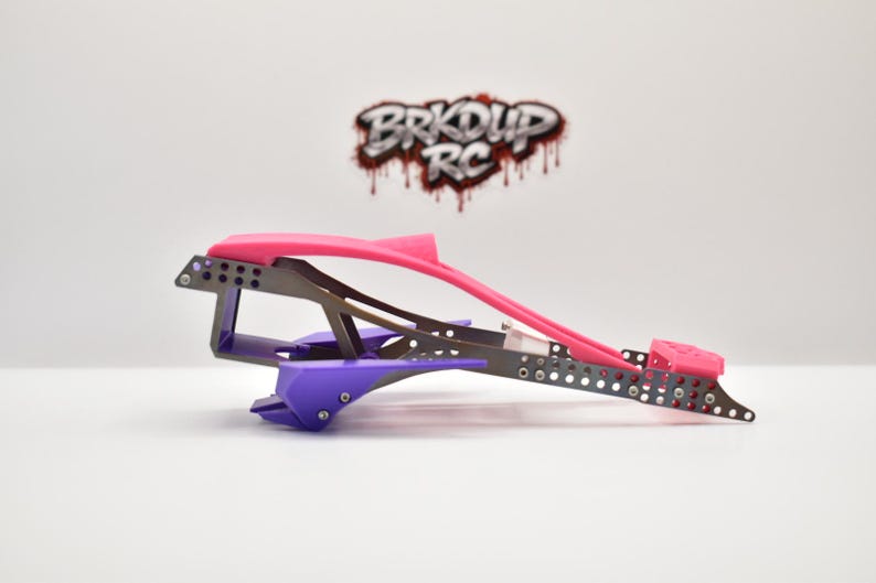 May include: A custom RC car chassis frame featuring a metallic grey perforated metal structure, a bright pink top section, and purple lower components. The logo BRKDUP RC is visible in the background in a stylized red and black font.