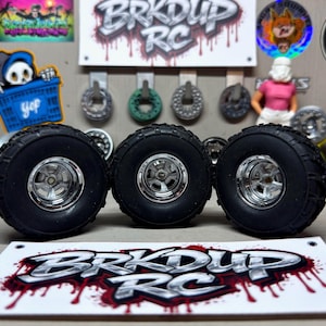 May include: Three black RC car tires with chrome rims are displayed. The tires have a rugged tread pattern. The background includes stickers, a sign that says "BRK DUP RC", and other RC car parts.