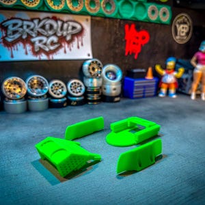 Op de afbeelding: Een afbeelding met RC-auto-onderdelen en accessoires. De afbeelding toont verschillende sets zilveren en zwarte wielen, samen met felgroene plastic componenten. Een bord met "BRKOUD RC" is zichtbaar op de achtergrond, samen met miniatuurbeeldjes.