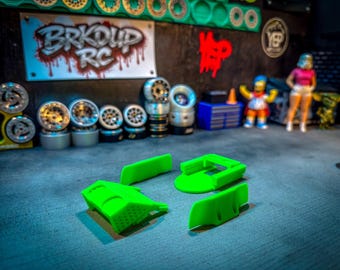 May include: A collection of RC car parts and accessories. The image features several sets of silver and black wheels, along with bright green plastic components. A sign reading "BRKOUD RC" is visible in the background, along with miniature figurines.
