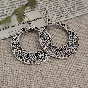 May include: A pair of circular silver-toned earrings featuring an intricate, embossed floral pattern with darkened recessed areas. Each earring is suspended from a simple silver-toned hook wire, showcasing a detailed, symmetrical design across the surface.