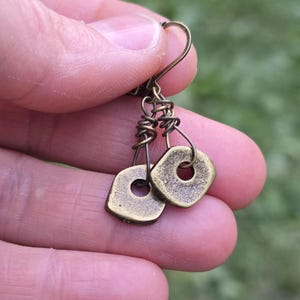 Può includere: Un paio di orecchini in ottone antico. Ogni orecchino presenta un piccolo pendente quadrato con un foro circolare al centro. I pendenti sono sospesi a un design avvolto in filo e attaccati a un gancio per l'orecchio. Gli orecchini hanno un aspetto vintage e artigianale.