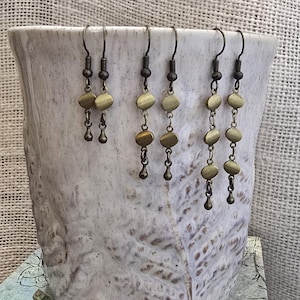 May include: A collection of antique brass-toned earrings. Each earring features a fishhook style ear wire, a small bead, three oval-shaped beads, and a teardrop charm. The earrings are displayed in a white ceramic container.