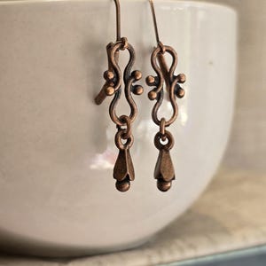May include: A pair of copper-colored earrings with an ornate design. Each earring features a curved top with small, round accents, a central looped element, and a teardrop-shaped dangle. The earrings are displayed against a white background.