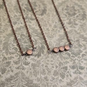 May include: Three copper-toned necklaces with delicate chains. One necklace features a single circular pendant, another has three, and the third has four. The necklaces are displayed on a patterned, muted green background.