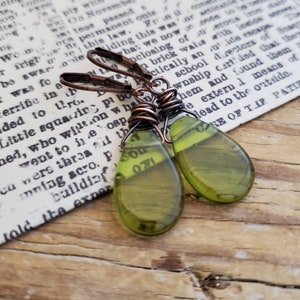 May include: A pair of handcrafted earrings with a vintage aesthetic. The teardrop-shaped pendants are olive green with visible text, suspended from copper-colored ear wires. The earrings are displayed on a background of aged paper and wood.