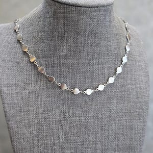 Puede incluir: Un collar plateado con una serie de pequeños colgantes circulares planos. Los colgantes están espaciados uniformemente y conectados por delicados eslabones, creando un diseño sencillo. El collar se exhibe en un maniquí gris.