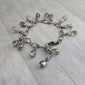 May include: A silver bracelet featuring a series of teardrop-shaped, clear and gray beads. The beads are suspended from a delicate chain with a lobster clasp closure. The bracelet is designed with a vintage or handcrafted aesthetic.