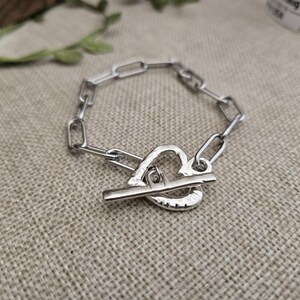 Pu&ograve; includere: Un braccialetto color argento con un design a catena e una chiusura a forma di cuore. Il braccialetto &egrave; realizzato in metallo lucido ed &egrave; esposto su una superficie di colore neutro. La chiusura &egrave; a levetta.
