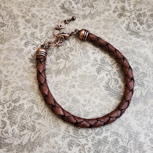 Puede incluir: Una pulsera de cuero trenzado marrón con detalles metálicos color cobre. La pulsera presenta un diseño tejido y un cierre ajustable. Los componentes metálicos incluyen extremos decorativos y una cadena para ajustar el tamaño.