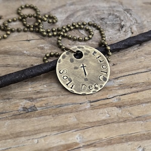 May include: A brass-colored pendant necklace with a hammered texture. The round pendant features the words "Soli Deo Gloria" and a cross symbol. The necklace has a ball chain and is displayed on a wooden surface.
