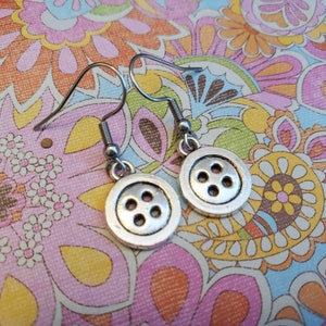 May include: A pair of silver button earrings with four holes on each button. The earrings are hanging from silver hooks.