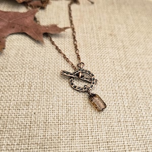 Op de afbeelding: Een koperkleurige ketting met een tuimelsluiting en een kleine, rechthoekige hanger. De ketting en sluiting hebben een getextureerd, gehamerd uiterlijk. De hanger heeft een doorschijnende, lichtbruine kleur. De ketting wordt gepresenteerd op een neutraal, getextureerd oppervlak.