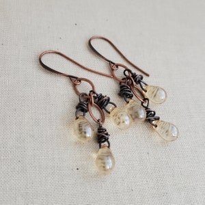 K&ouml;nnte beinhalten: Ein Paar H&auml;ngeohrringe mit Kupferdraht, der um klare Glas-Tropfenperlen gewickelt ist. Die Ohrringe haben einen rustikalen, handgefertigten Look.