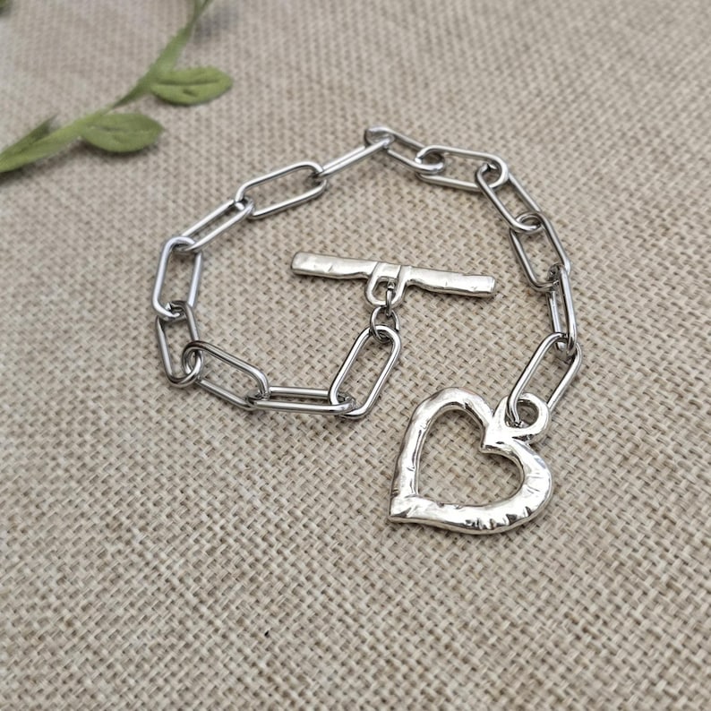 Pu&ograve; includere: Un braccialetto color argento con un ciondolo a forma di cuore e una chiusura a leva. Il braccialetto presenta un design a maglie di catena. Il ciondolo a forma di cuore ha una superficie strutturata.