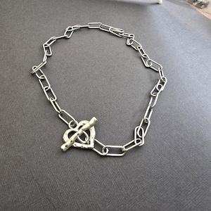 Pu&ograve; includere: Collana color argento con chiusura a forma di cuore. La catena &egrave; composta da maglie rettangolari allungate. La chiusura a cuore ha una superficie strutturata e una chiusura a barra. Il gioiello &egrave; presentato su una superficie grigia.