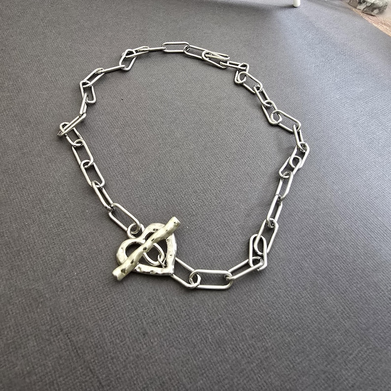 Pu&ograve; includere: Collana color argento con chiusura a forma di cuore. La catena &egrave; composta da maglie rettangolari allungate. La chiusura a cuore ha una superficie strutturata e una chiusura a barra. Il gioiello &egrave; presentato su una superficie grigia.