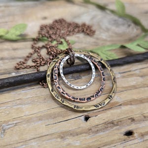 Puede incluir: Un collar en capas con tres anillos metálicos texturizados en tonos plata, cobre y latón. El colgante cuelga de una delicada cadena color cobre, sobre una superficie de madera rústica. El diseño tiene una estética bohemia y terrosa.