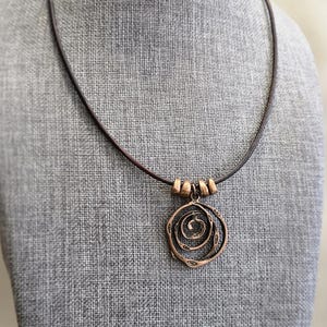 May include: A copper-toned pendant necklace with a spiral design. The pendant is suspended from a dark brown leather cord, with three small copper beads near the clasp. The necklace is displayed on a grey mannequin.