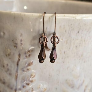 May include: A pair of copper-coloured earrings. Each earring has a teardrop-shaped pendant with a small ball at the bottom, suspended from a simple hook. The earrings are set against a blurred, off-white background.