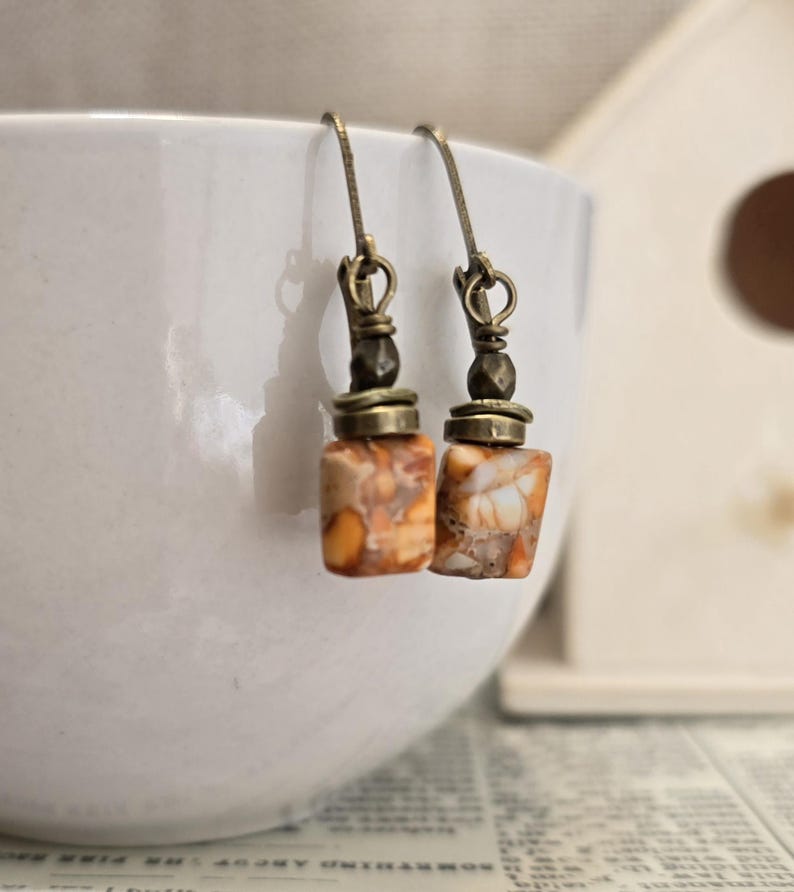 May include: A pair of handcrafted earrings featuring rectangular, speckled orange and white beads. The earrings have antique brass-colored hooks and decorative elements. The beads have a unique, natural stone appearance, adding a touch of earthy elegance to the jewelry.