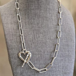 May include: A silver chain necklace with a heart-shaped clasp. The chain is made of linked oval shapes.