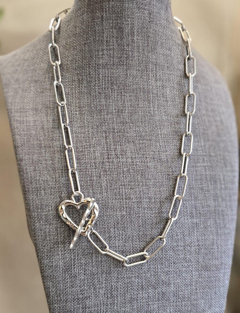 Pu&ograve; includere: Collana color argento con un design a catena a maglie grandi. La collana presenta un ciondolo a forma di cuore con chiusura a barra. Il gioiello &egrave; esposto su un manichino grigio, che mette in risalto la lunghezza e lo stile della collana.