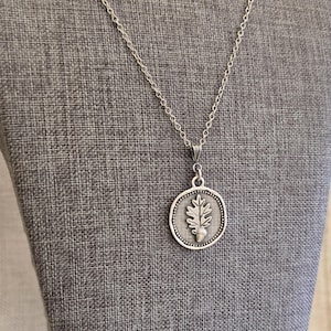 May include: Silver-toned necklace featuring a circular pendant with an oak leaf and acorn design. The pendant is detailed with a dotted border and hangs from a delicate chain. The necklace is displayed against a gray fabric background.