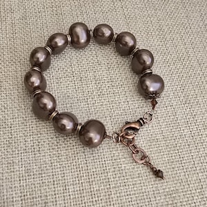 May include: A bracelet featuring round, dark brown, lustrous beads separated by small metallic spacers. The piece includes a copper-toned lobster claw clasp and an adjustable chain extension finished with a small, dark, faceted bead at the end.