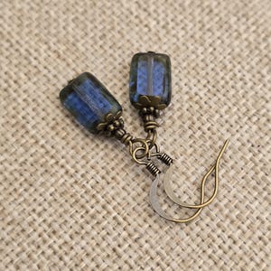 May include: A pair of handcrafted earrings featuring rectangular blue and green glass beads. The beads are suspended from antique brass-colored metal findings and ear wires. The earrings are displayed on a textured, neutral-toned surface.