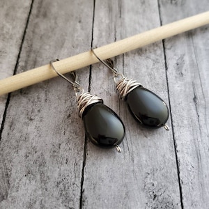 Pode incluir: Um par de brincos pretos em forma de gota. Cada brinco apresenta uma pedra preta polida, envolta em fio prateado e um fecho de alavanca. Os brincos são exibidos em um pino de madeira sobre um fundo de grão de madeira cinza.