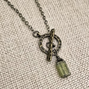 Puede incluir: Un collar de estilo vintage con un cierre de palanca y un pequeño colgante rectangular de piedra verde. La cadena y el cierre tienen un acabado bronce, y el colgante es de color verde pálido. El collar se muestra sobre una superficie texturizada de color neutro.