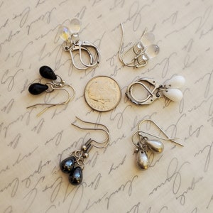 May include: Assortment of six pairs of dangle earrings with silver-toned hooks. The earrings feature teardrop-shaped stones in black, white, and iridescent.