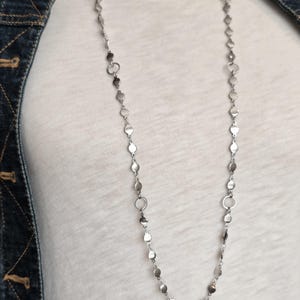 Puede incluir: Un collar largo de tono plateado que presenta una cadena delicada intercalada con pequeños discos metálicos planos y circulares y anillos abiertos más grandes ocasionales, creando un patrón repetitivo que cuelga verticalmente sobre un fondo de tela blanca.