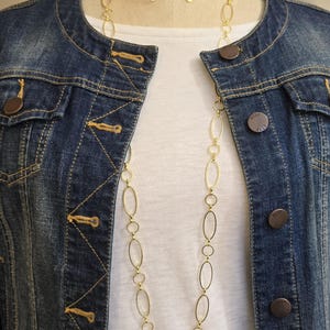 May include: A gold-toned necklace and matching earrings with an oval link design. The jewelry is displayed on a mannequin, layered over a white top and a blue denim jacket with gold stitching and buttons.