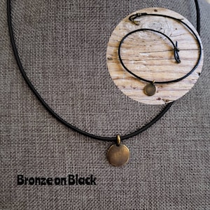 以下が含まれることがあります： 黒い革ひもでできたシンプルなネックレスで、丸いブロンズのペンダントが付いています。ネックレスの下に「Bronze on Black」という文字が見えます。